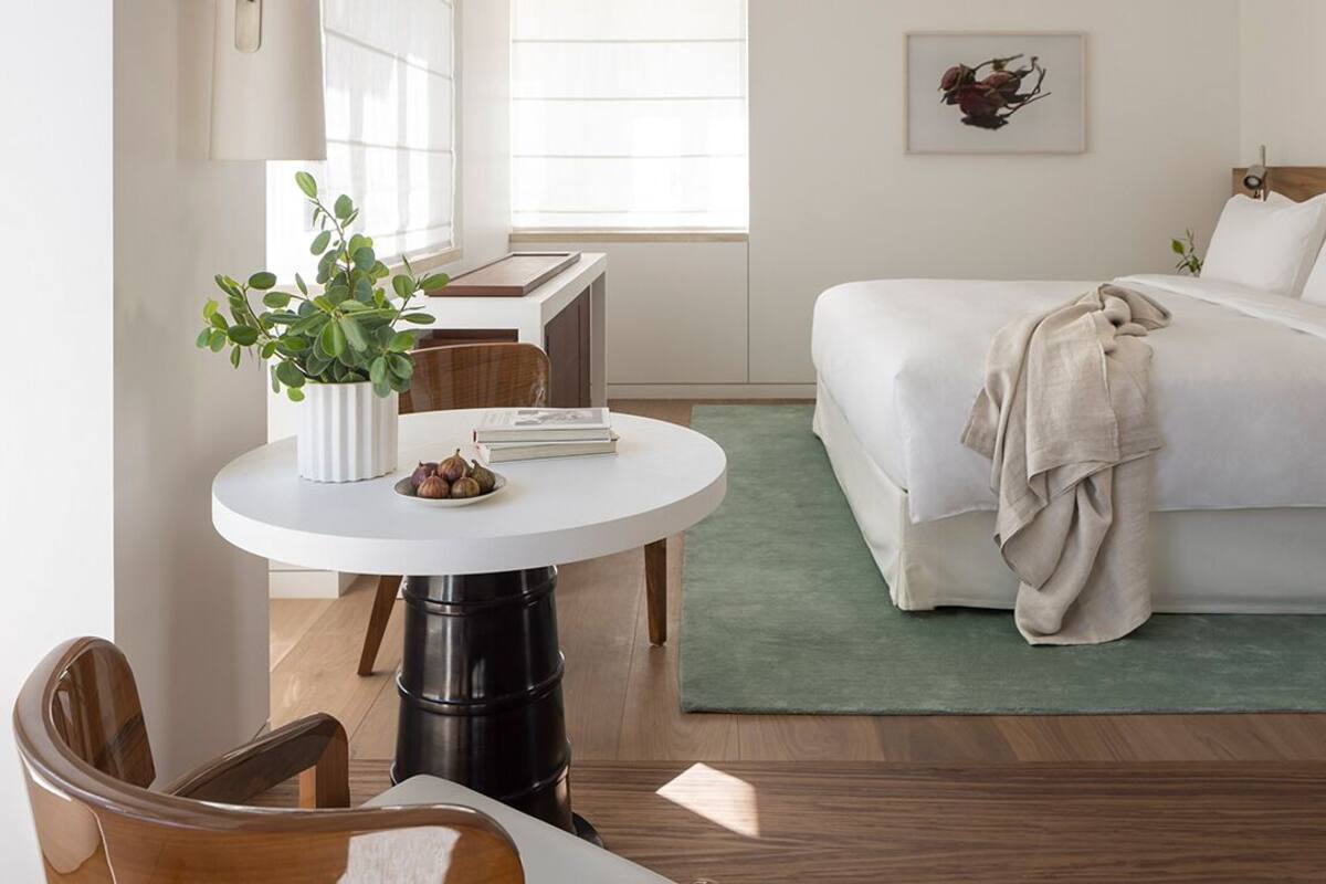 Una habitación de hotel con una cama, una mesita redonda con una planta, una silla y un escritorio con una lámpara.