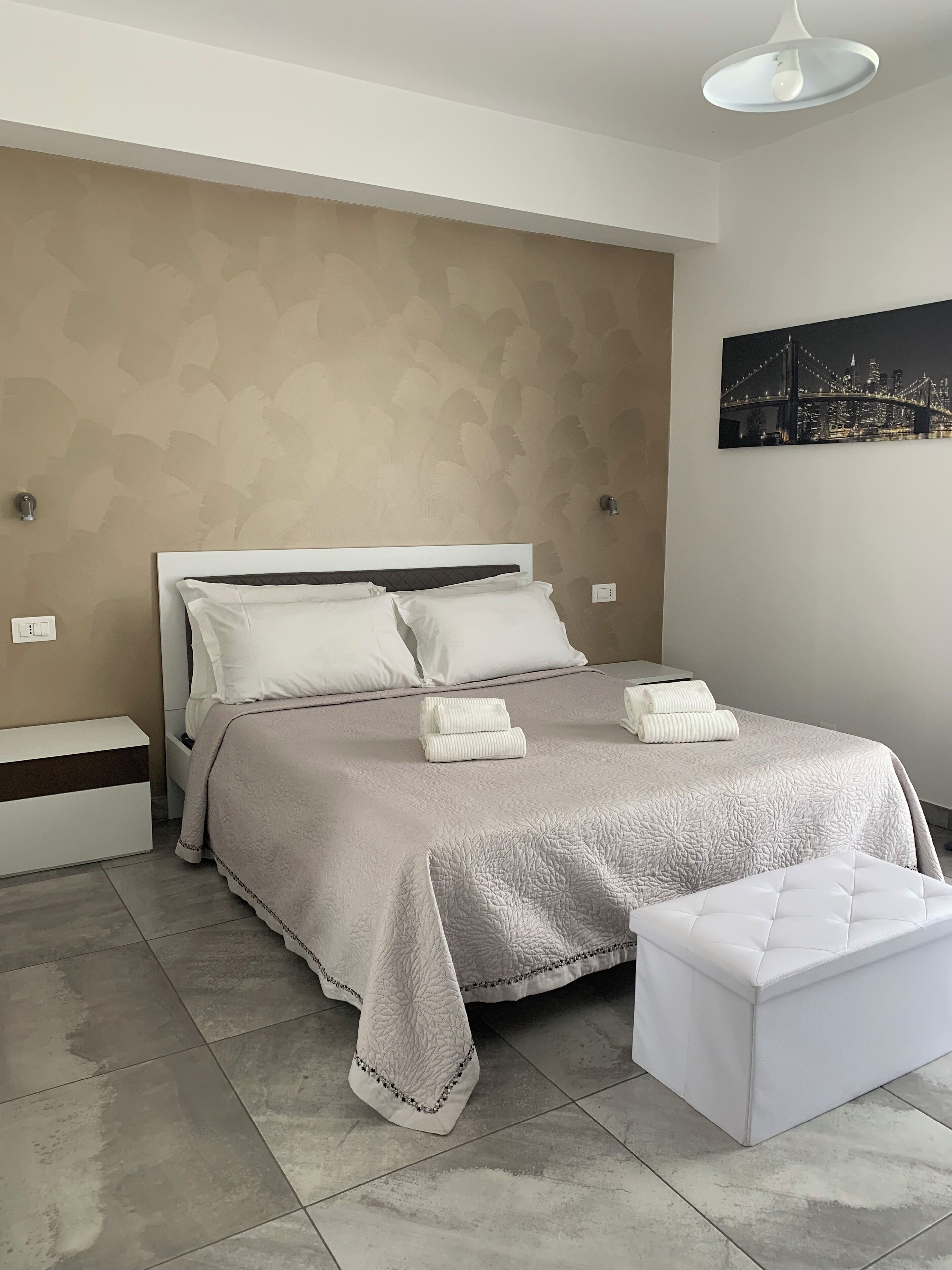 A neatly arranged bedroom with a bed, bedside tables, a bench, and a cityscape artwork on the wall.