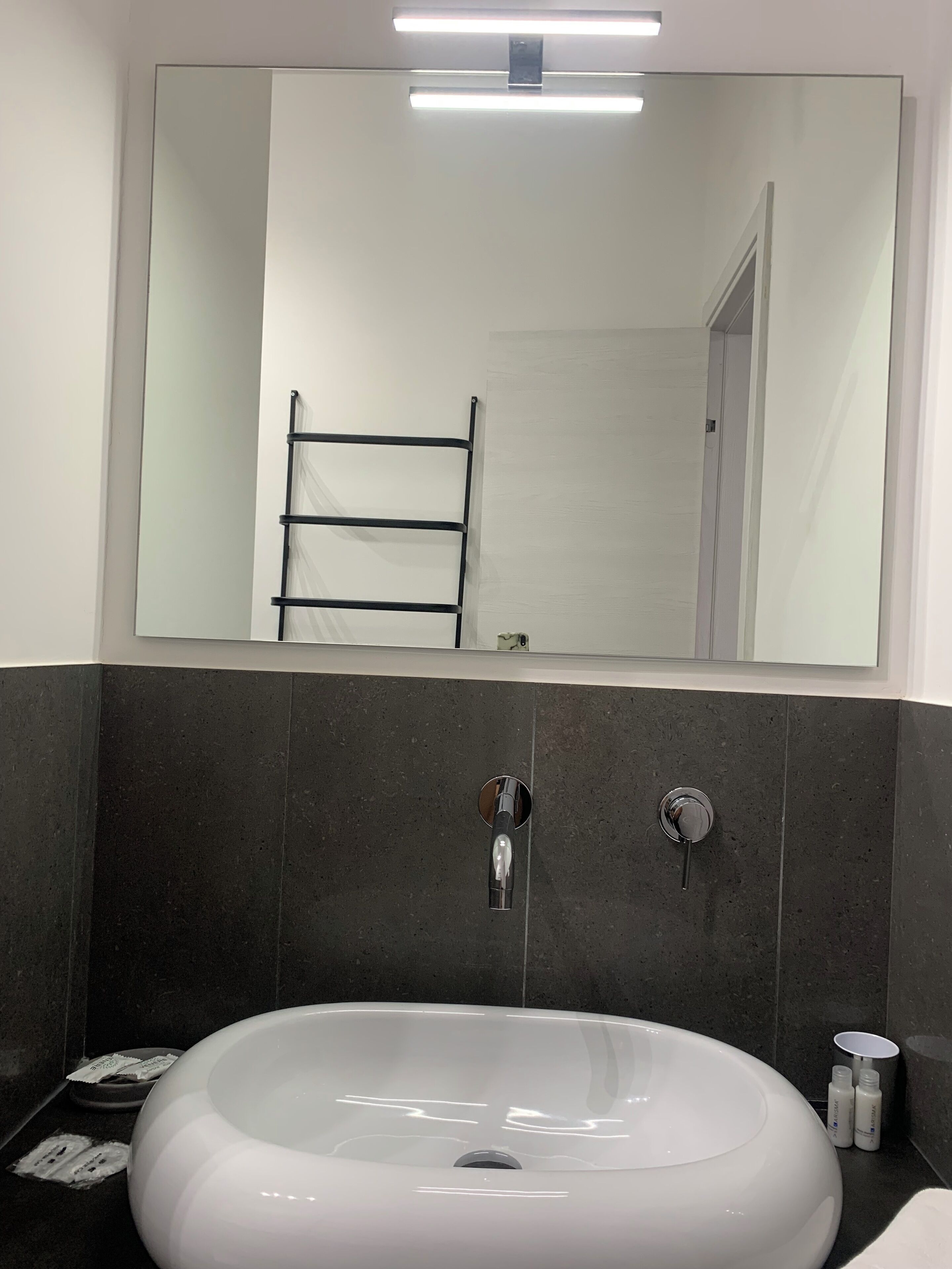 A modern bathroom with a large mirror, a white sink, dark tiled walls, and a towel rack.