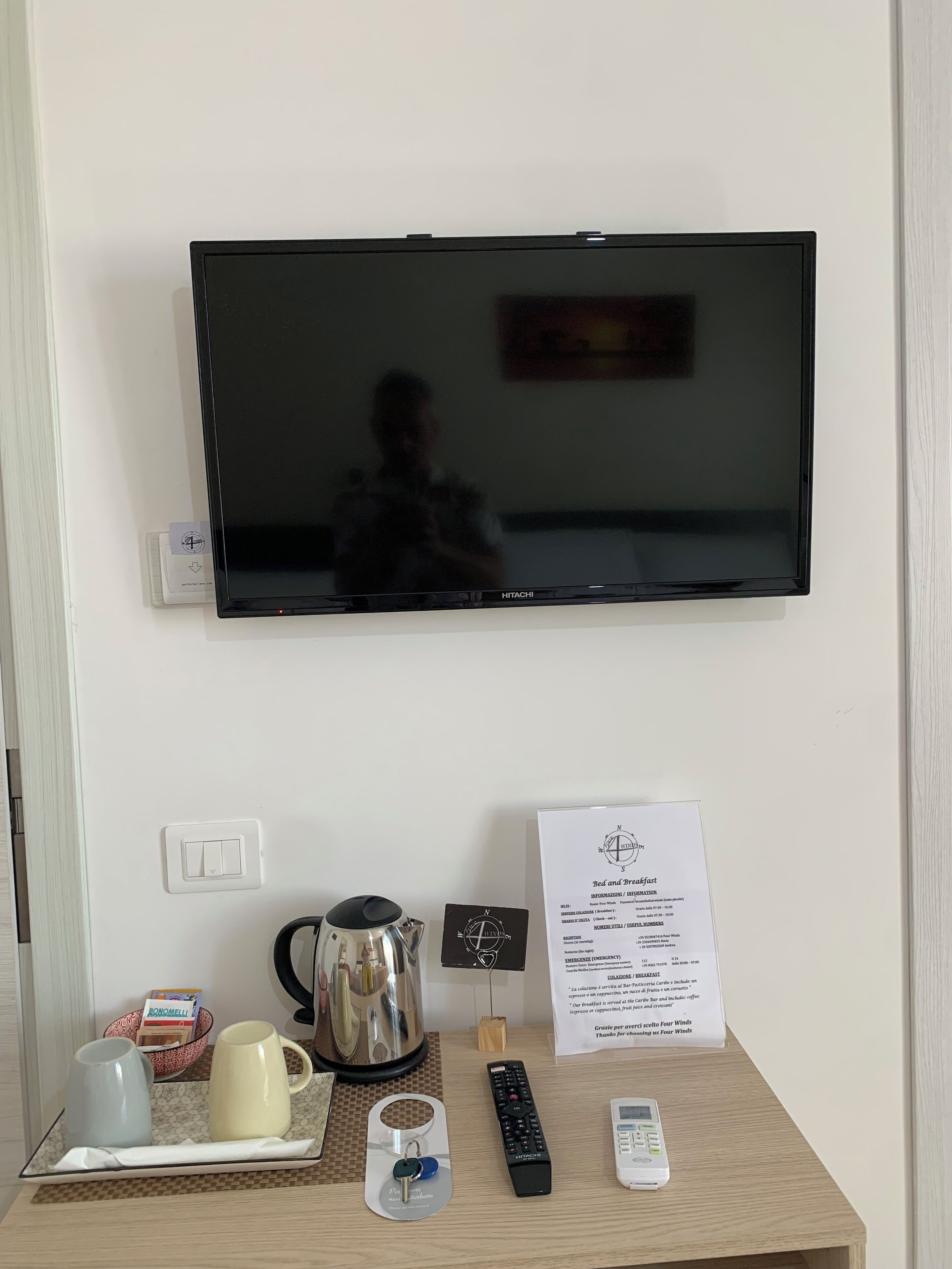 Habitación de hotel con televisor de pantalla plana en la pared, mesita de noche con hervidor, tazas, control remoto, carta y un reloj pequeño.