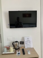 Habitación de hotel con televisor de pantalla plana en la pared, mesita de noche con hervidor, tazas, control remoto, carta y un reloj pequeño.