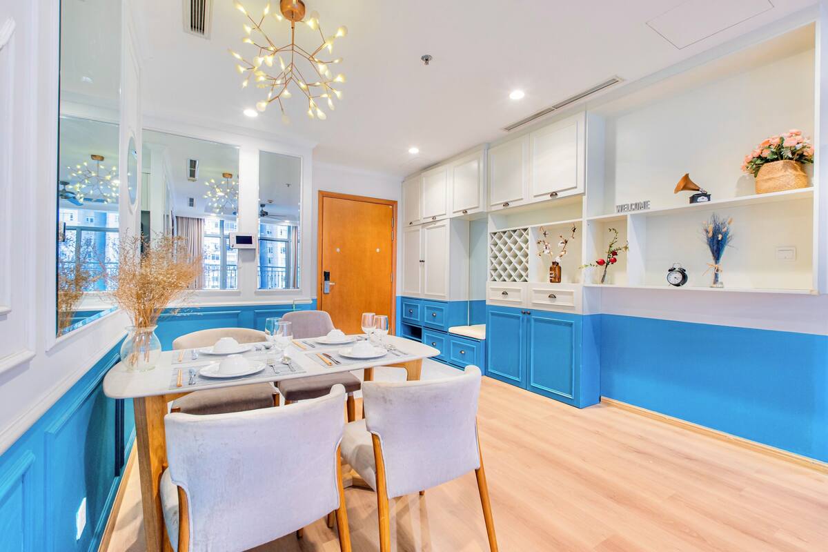 Une cuisine moderne avec des meubles de couleur bleue et blanche, une table à manger pour quatre personnes et un vase décoratif posé sur le rebord de la fenêtre.