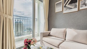 Een moderne woonkamer met een beige bank, een tafel met een tijdschrift en een bloemenvaas. Een groot raam biedt uitzicht op een stadsgezicht en een balkon met een hekwerk.