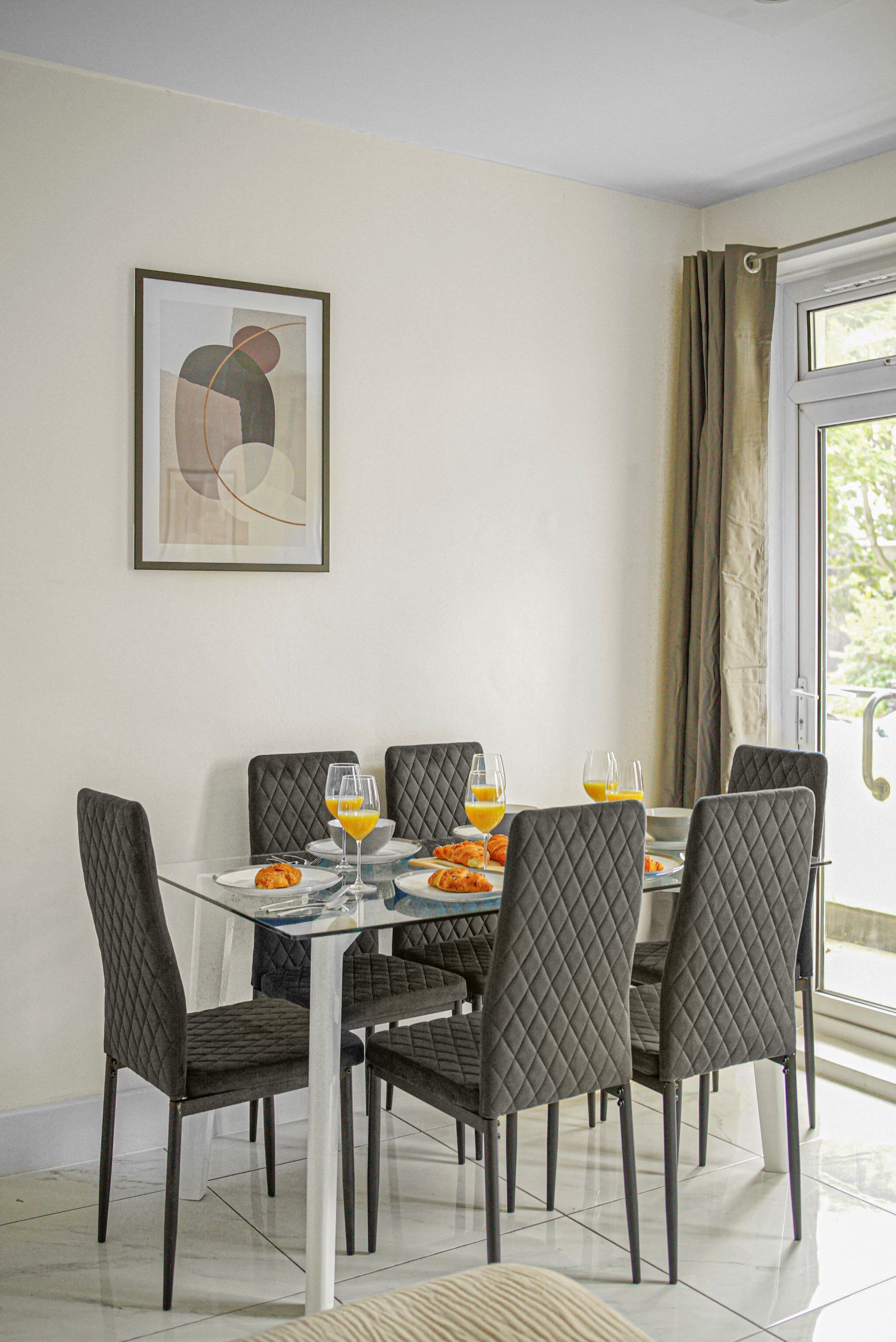 A dining area with a glass table and cushioned chairs, a framed abstract painting on the wall, and a view through a window with curtains.