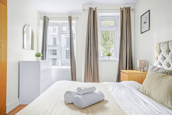 Habitación de hotel con cama, cómoda, mesita de noche, espejo y ventana con cortinas.