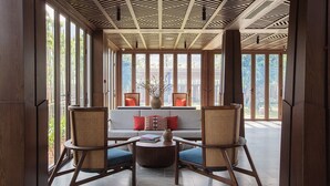 Une pièce avec un plafond en bois, de grandes fenêtres, un canapé, deux fauteuils en osier et une table ronde sur laquelle sont posés des livres.