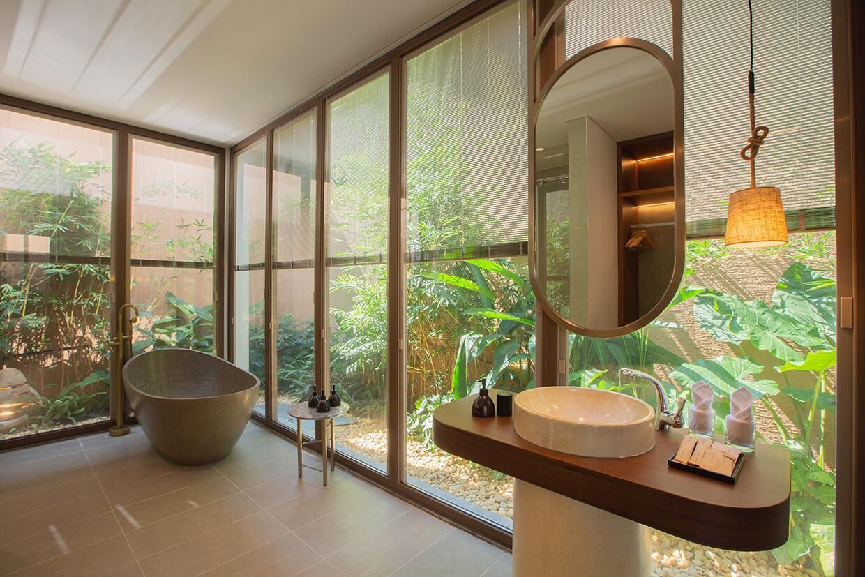 A modern bathroom with a large bowl sink, a round mirror, and a view of lush greenery through large windows.