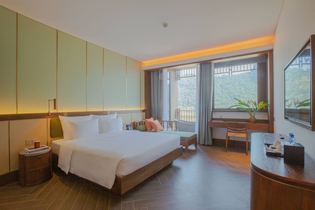 Una habitació d'hotel amb un llit gran, una taula i una cadira. Hi ha una finestra amb vista a la vegetació a l'exterior.