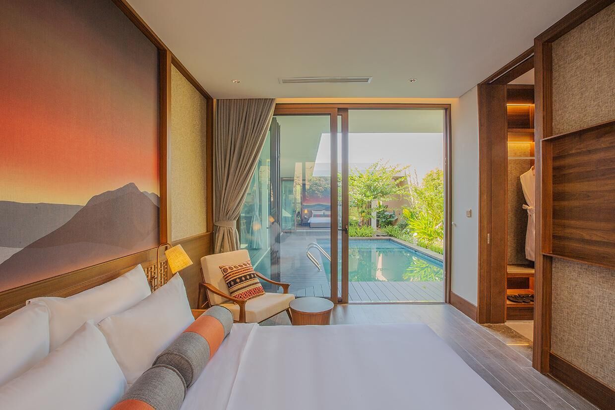 Habitación de hotel con cama, una silla, una mesita, un cuadro y vistas a una piscina y zonas verdes.