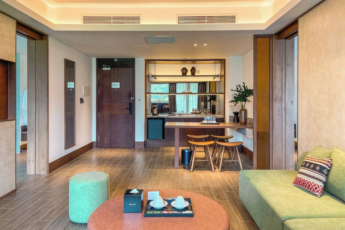 Habitació d'hotel moderna amb sòl de fusta, un sofà verd, una taula de cafè rodona i una zona de cuina amb barra i seients.