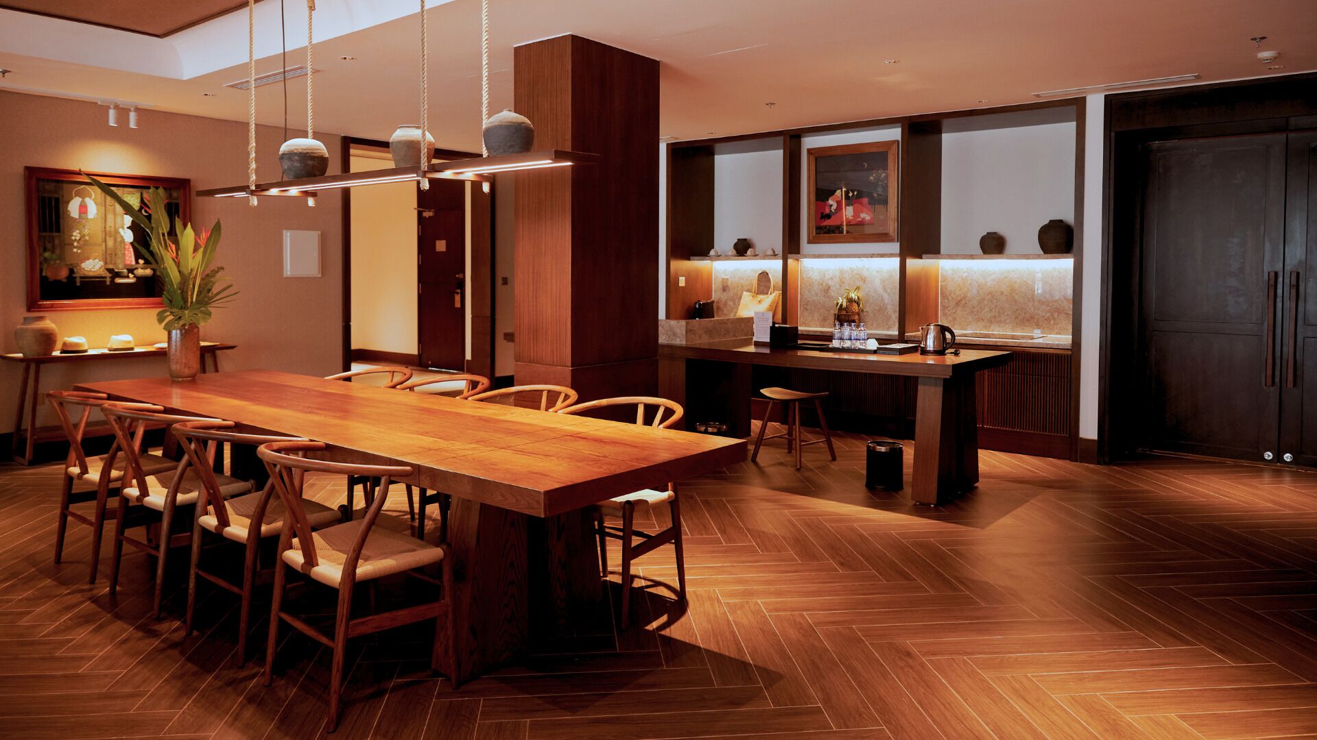 Un comedor moderno con una mesa y sillas de madera, una lámpara colgante y una zona de cocina con mostrador y taburetes.