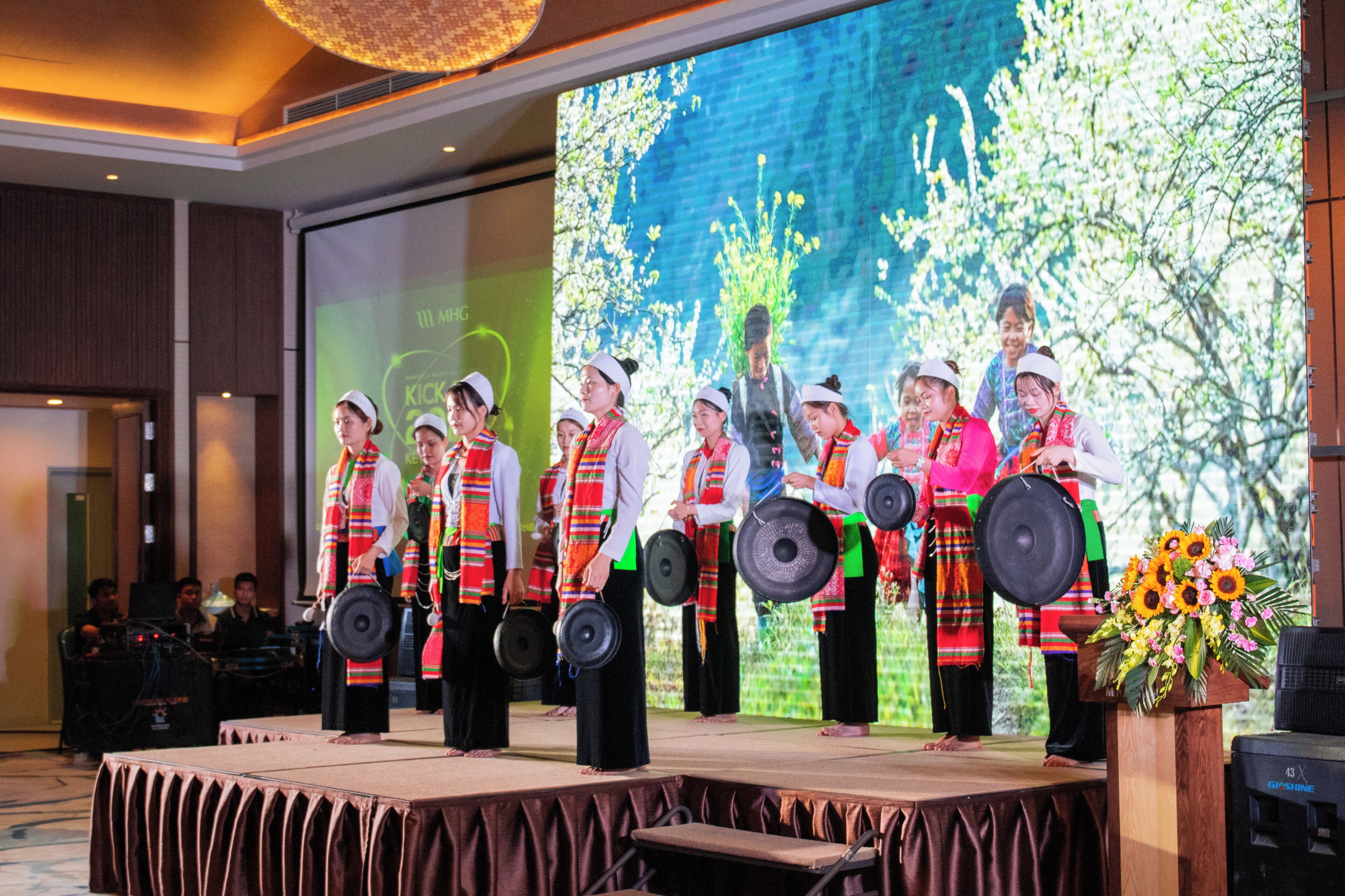 Un escenario con artistas vestidos con atuendos tradicionales, una pantalla grande al fondo y una decoración floral.