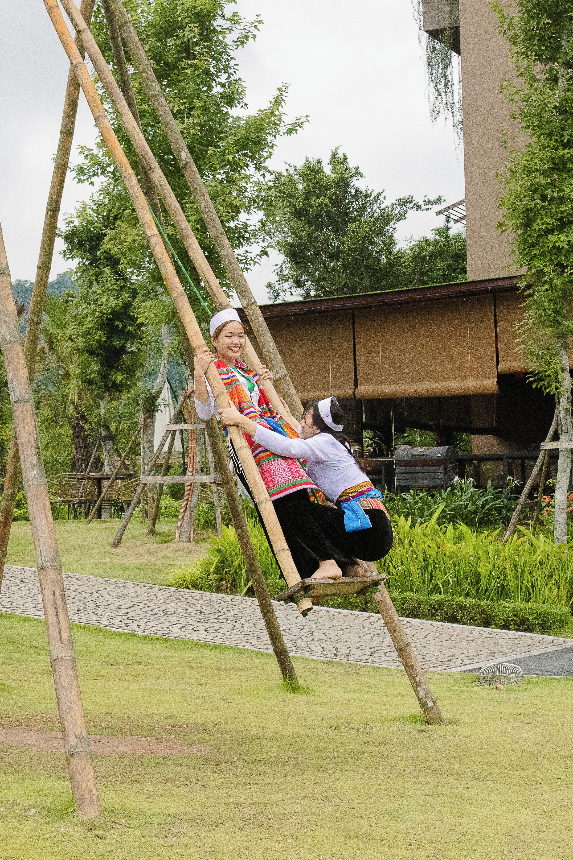 A person using a bamboo swing in a garden area with a building in the background.