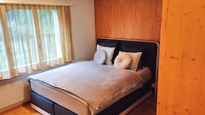 Un lit simple avec une tête de lit sombre, deux oreillers et une couverture beige, dans une pièce aux murs lambrissés et dotée d’une fenêtre ornée de rideaux légers.