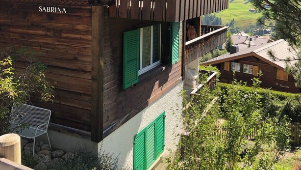 Un chalet en bois doté d’un balcon, de volets verts et offrant une vue sur un paysage montagneux.
