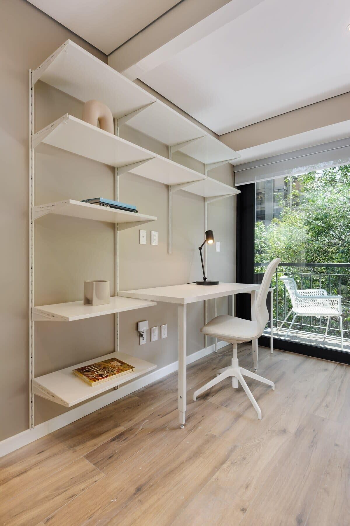A modern room with a white desk, shelves, and a chair. There is a window with a view of greenery outside.