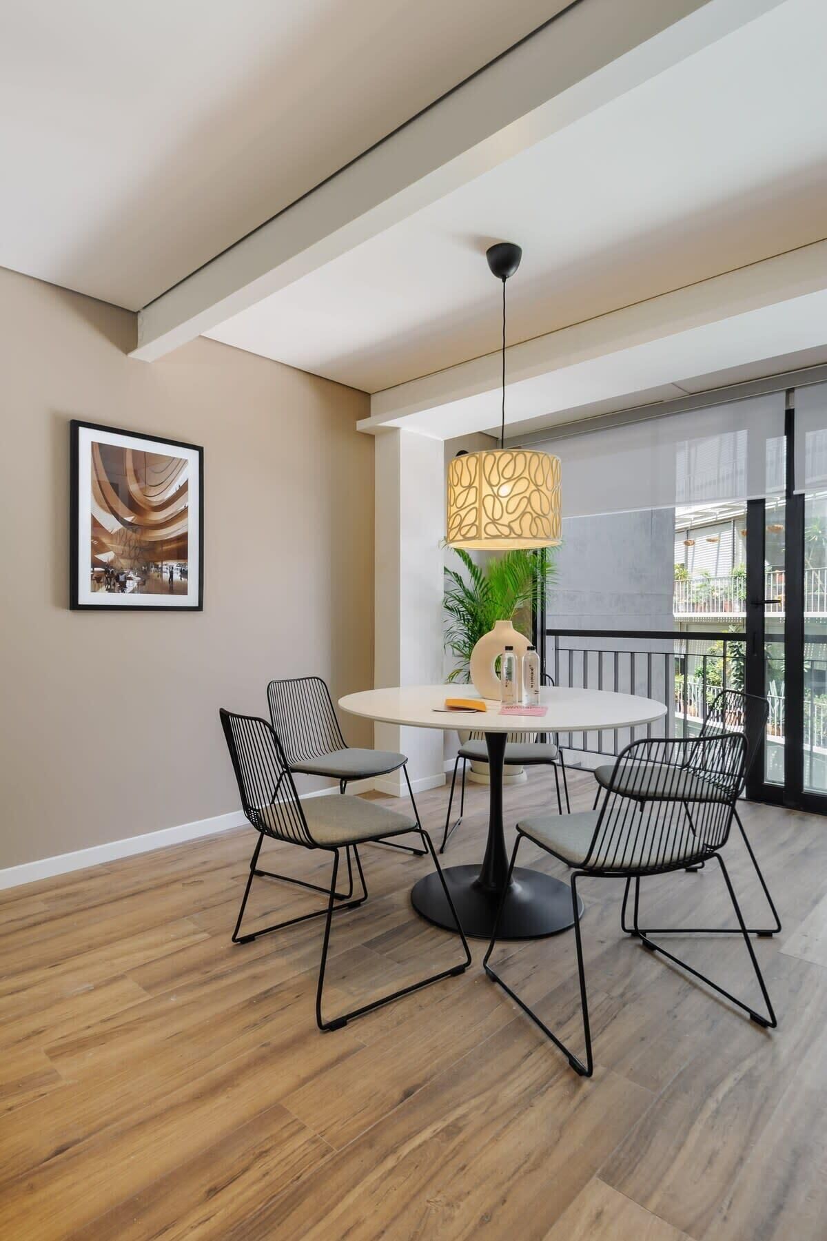 A modern dining area with a round table, four black chairs, a pendant light, and a framed picture on the wall.