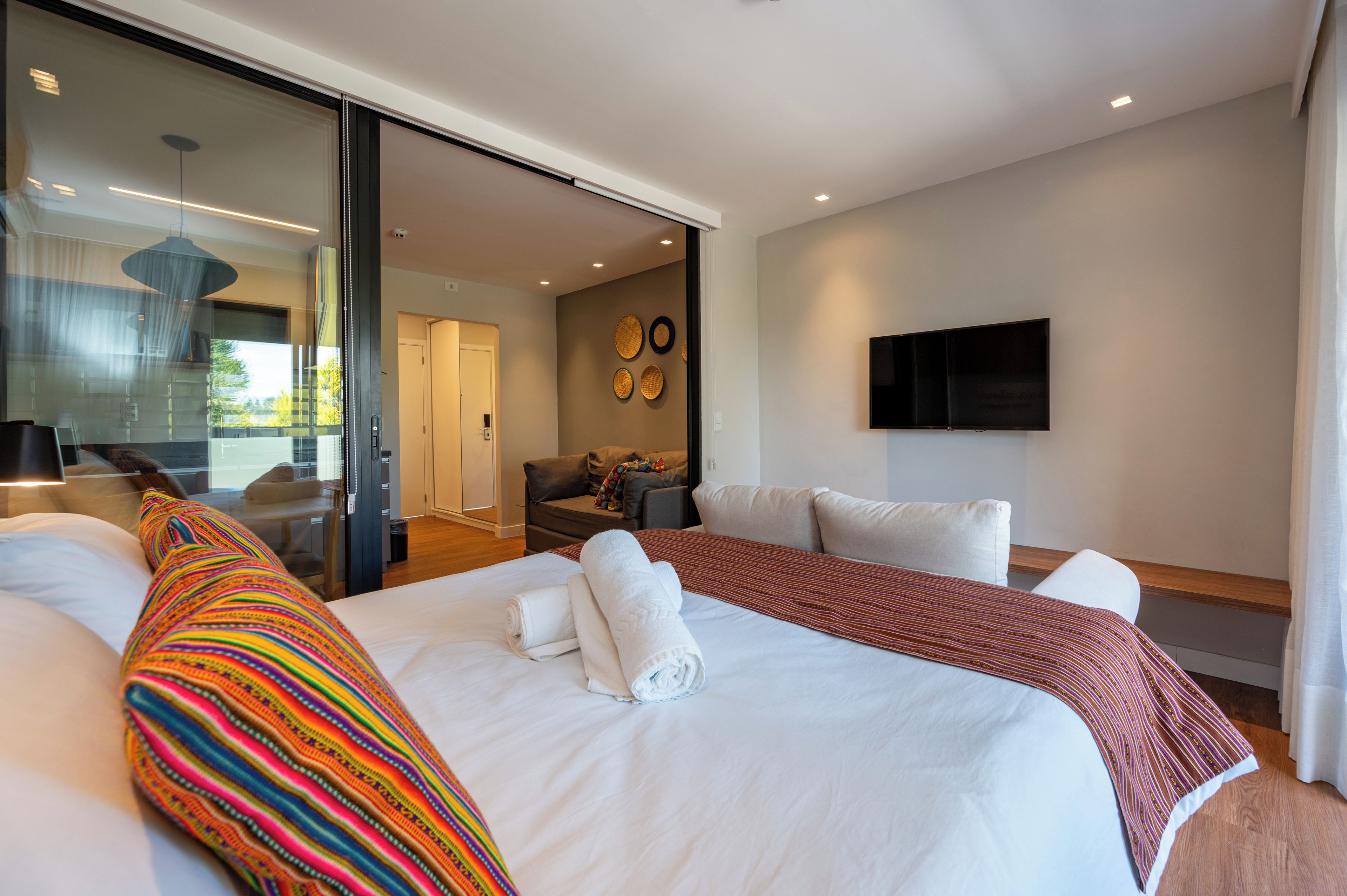 Quarto de hotel moderno com uma cama grande, um sofá, uma televisão e uma porta de vidro deslizante que dá para uma varanda.