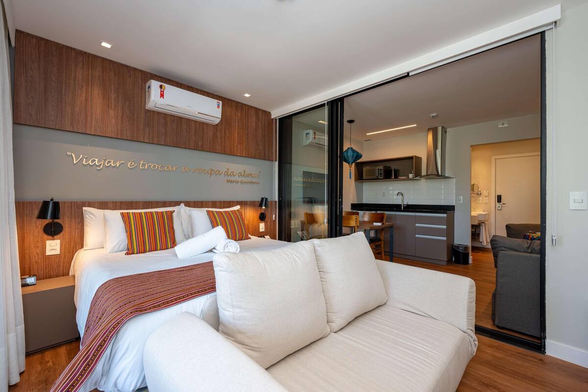 A modern hotel room with a bed, a sofa, a kitchenette, and a sliding glass door to the living area.