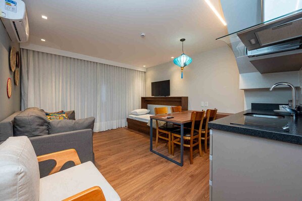 A modern hotel room with a sofa, a dining table with chairs, a bed, a TV, a kitchen area with a sink, and a ceiling-mounted air conditioning unit.