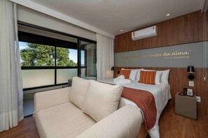A modern hotel room with a large bed, a sofa, a wooden accent wall, and a view of greenery outside. - Wanderlust Experience Hotel, BW Signature (Campos do Jordão)