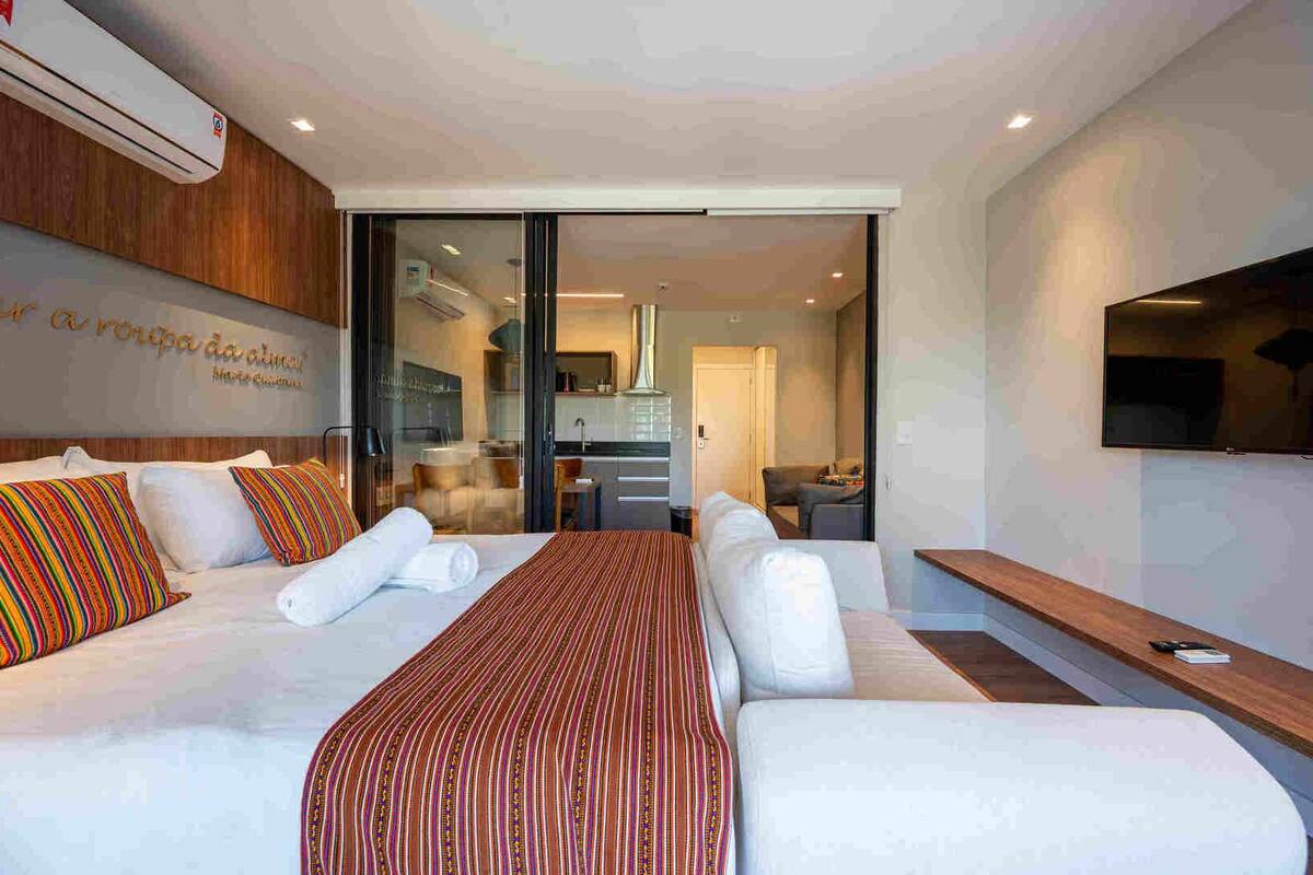 A modern hotel room with a large bed, a TV mounted on the wall, and a sliding glass door leading to a kitchen area.