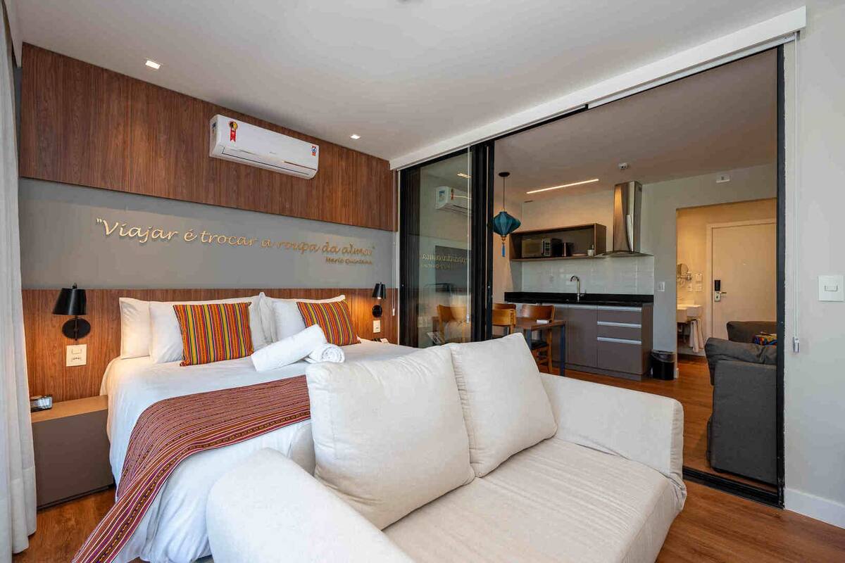 A modern hotel room with a bed, a sofa, a kitchen area, and a sliding glass door leading to a balcony.