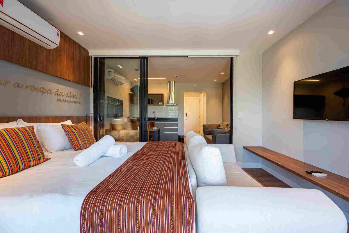 A modern hotel room with a large bed, a TV mounted on the wall, and a sliding glass door leading to a kitchen area.