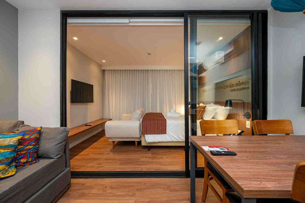 A modern hotel room with a bed, a sofa, a dining table, and a TV.