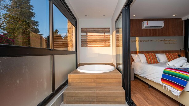 A modern bathroom with a freestanding bathtub, a wooden bench, and a bed with a colorful blanket. - Wanderlust Experience Hotel, BW Signature (Campos do Jordão)