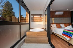 A modern bathroom with a freestanding bathtub, a wooden bench, and a bed with a colorful blanket.
