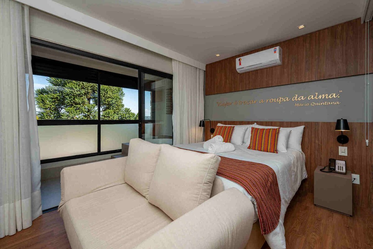 A modern hotel room with a large bed, a sofa, a wooden accent wall, and a view of greenery outside.
