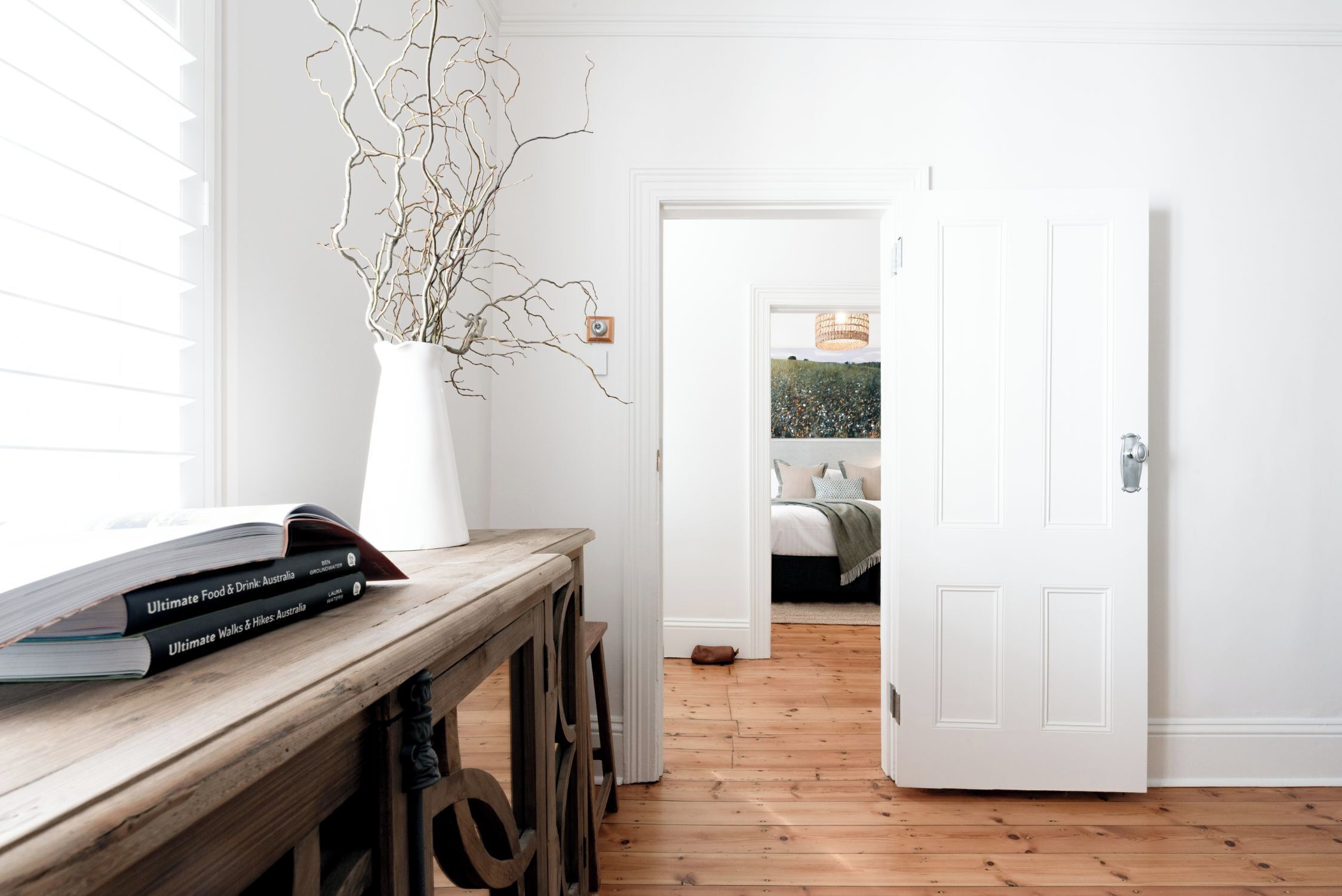 A room with a wooden floor, a white door, and a window with blinds. There is a white vase with branches on a wooden table.