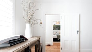 Ein Zimmer mit Holzboden, einer weißen Tür und einem Fenster mit Jalousien. Auf einem Holztisch steht eine weiße Vase mit Zweigen.
