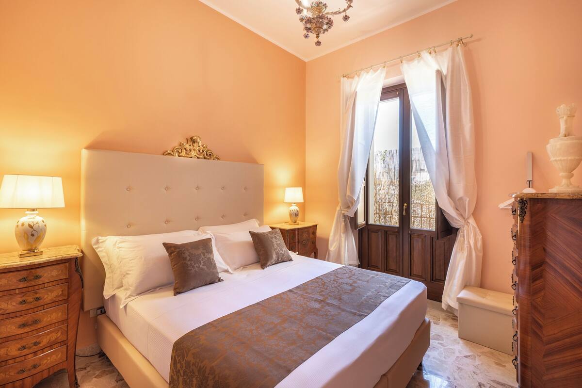A bedroom with a large bed, two bedside tables, a nightstand, a wooden dresser, and a window with curtains.