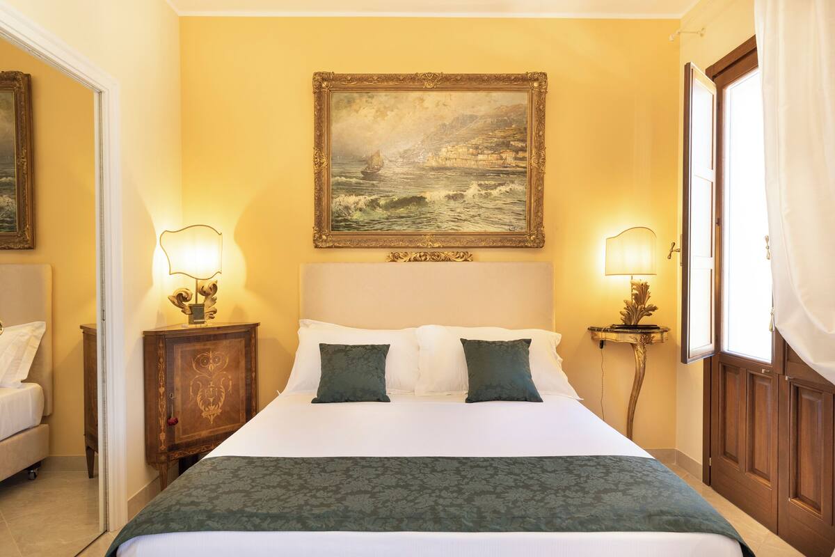A hotel room with a bed, two bedside lamps, a painting on the wall, and a door with curtains.