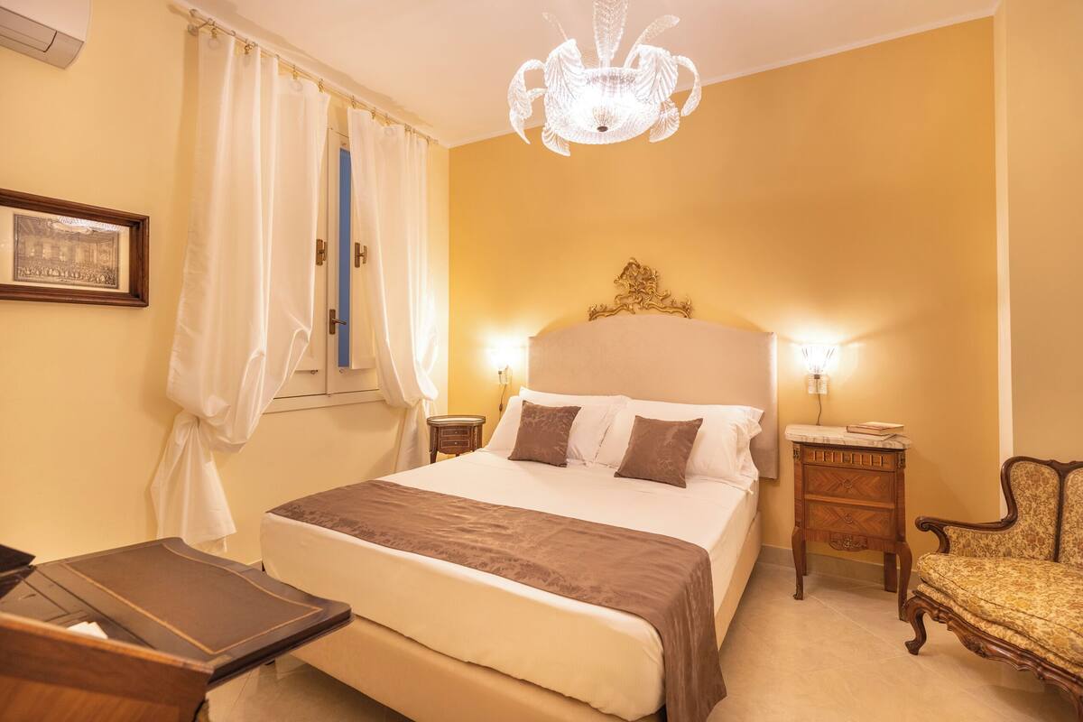 A hotel room with a large bed, a wooden nightstand, a chair, and a chandelier.