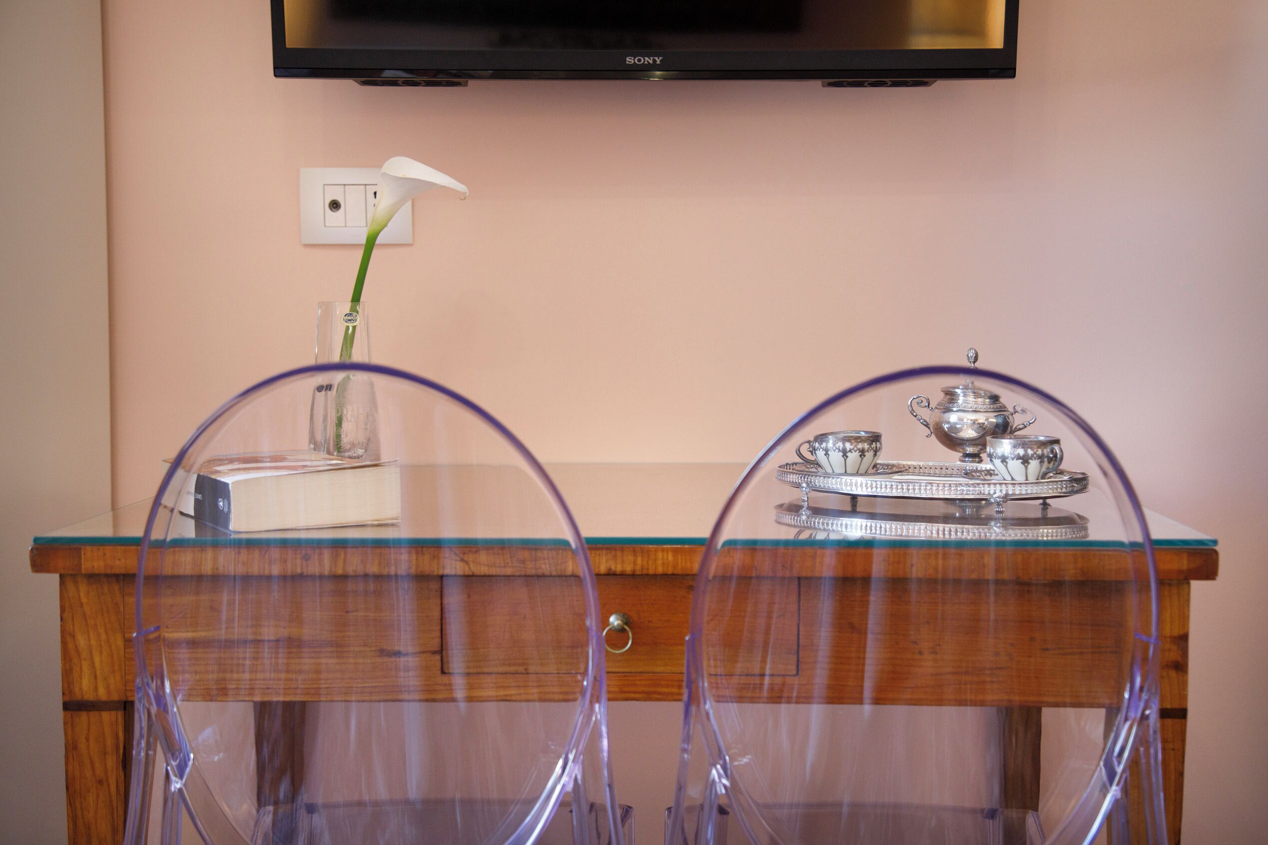 Un bureau en bois surmonté d’une plaque de verre, deux chaises transparentes, un vase avec une fleur blanche et un plateau en argent contenant des tasses et des soucoupes.
