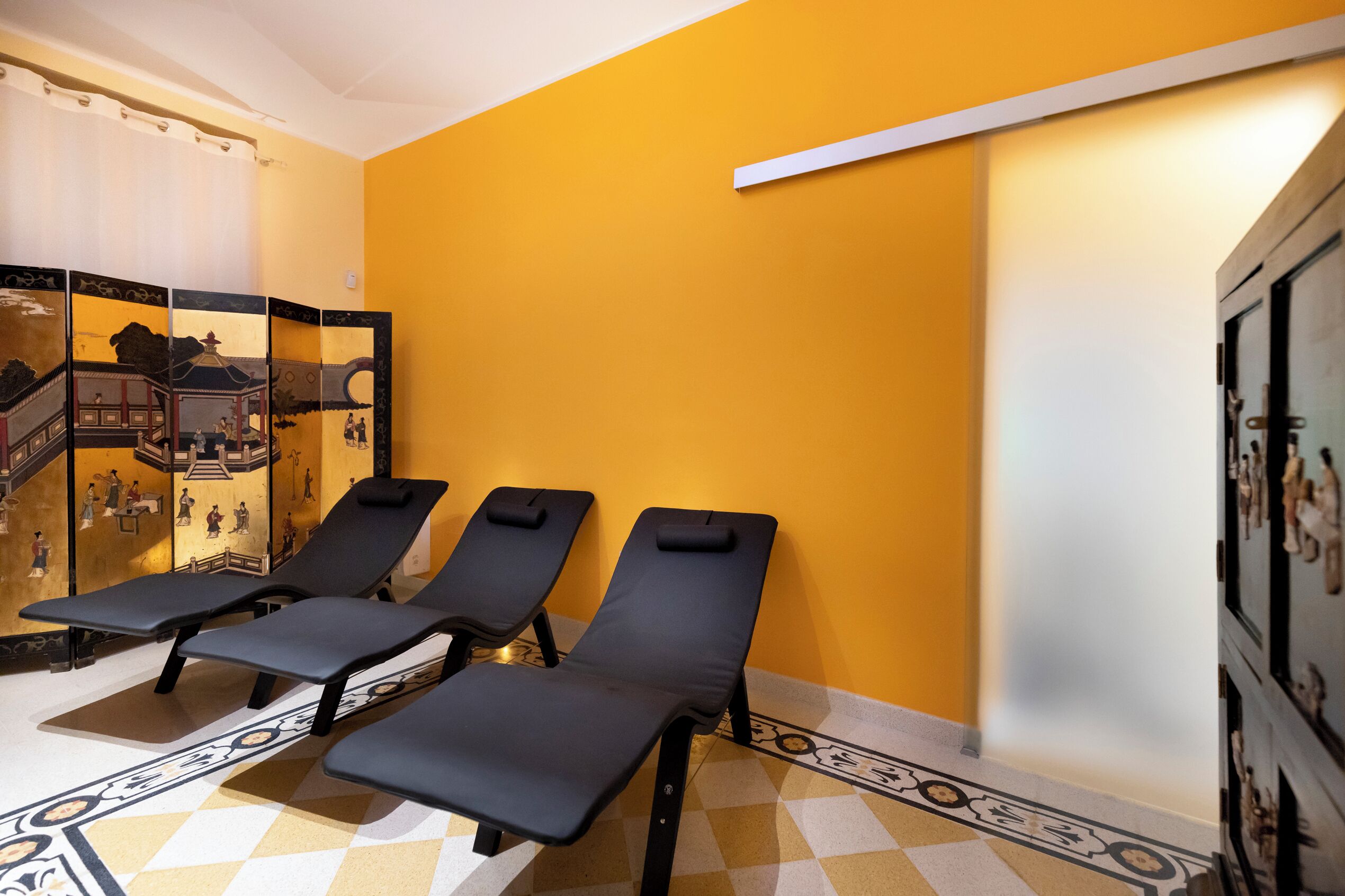 A room with four black sun loungers against a yellow wall, a decorative screen with traditional artwork, and a patterned floor.
