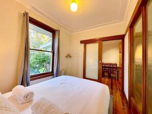 A hotel room with a bed, a window with curtains, a wooden floor, and a sliding door leading to a dining area.