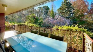 Um pátio externo com mesa e cadeiras, cercado por um portão decorativo e árvores.