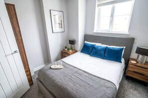 Un dormitorio con cama, mesitas de noche, una ventana y una puerta.