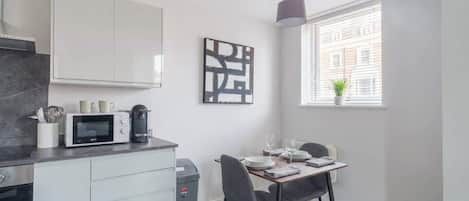 A modern kitchen with a microwave, coffee maker, and utensils on the counter. There is a dining table with chairs and a window with blinds.