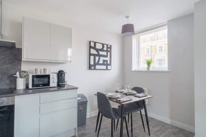 A modern kitchen with a microwave, coffee maker, and utensils on the counter. There is a dining table with chairs and a window with blinds.