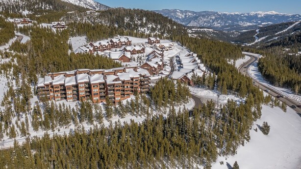 Ein Skigebiet mit mehreren Berghütten, einer kurvenreichen Straße und schneebedeckten Bergen im Hintergrund.