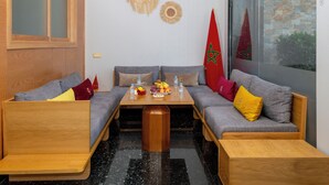 Ein modernes Zimmer mit einer Sitzecke mit Holzbank, einem Tisch mit Früchten und Wasserflaschen, marokkanischen Flaggen und Blick durch ein Fenster.