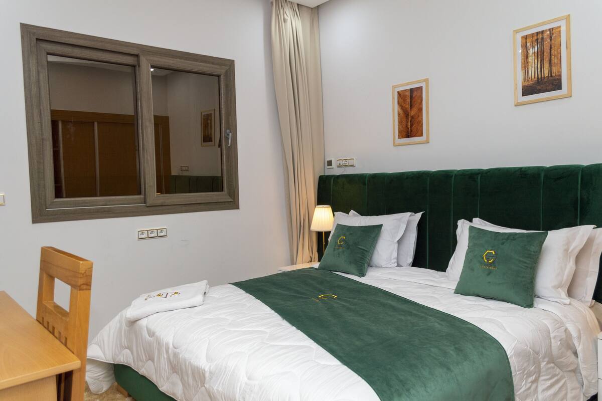 Un lit bien fait, recouvert d’une couverture verte assortie aux coussins, une chaise en bois, une table et une fenêtre avec des rideaux.