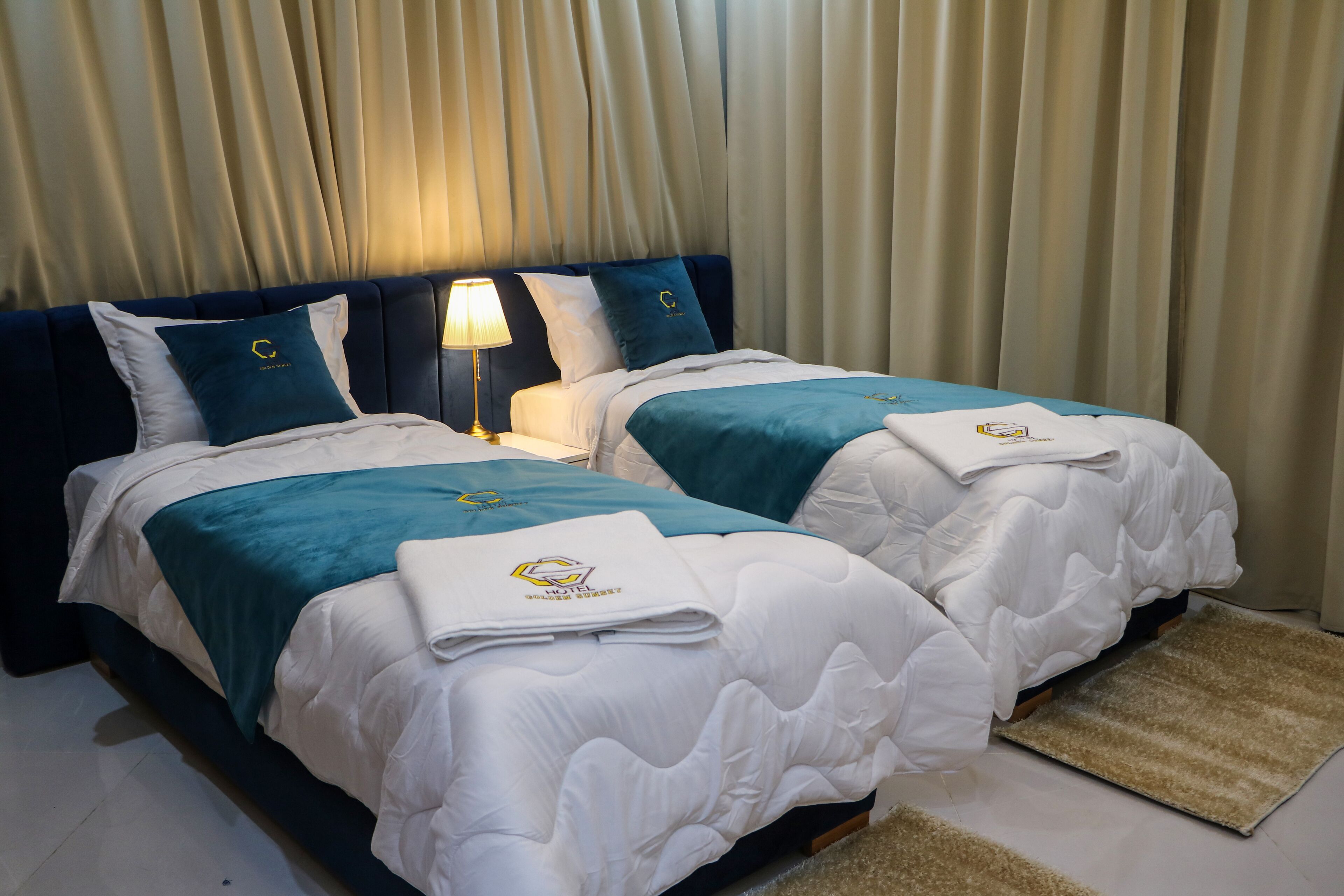A hotel room with two beds, each with a teal bedspread and white bedding. There is a lamp on one side of the beds, and a beige curtain in the background.