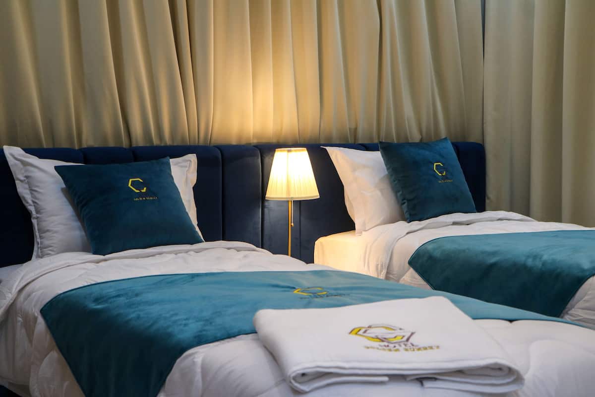Une chambre d’hôtel avec deux lits, chacun recouvert d’une couvre-lit et doté d’oreillers de couleur turquoise, avec une tête de lit blanche et une lampe placée entre les deux lits.