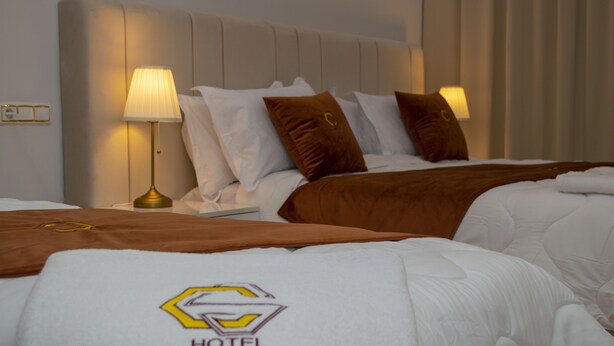 Ein Hotelzimmer mit zwei Betten, jeweils mit brauner Kopfteil und weißer Bettwäsche. Zwei Nachttischlampen und eine sichtbare Steckdose an der Wand.
